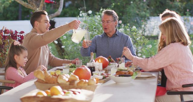 Family gathered around outdoor picnic table for meal on sunny day. Glasses raised, enjoying lemonade, fresh bread and grilled foods. Vibrant garden backdrop with flowers and greenery creates warm, inviting atmosphere. Ideal for concepts around family bonding, outdoor activities, summer gatherings and healthy living.