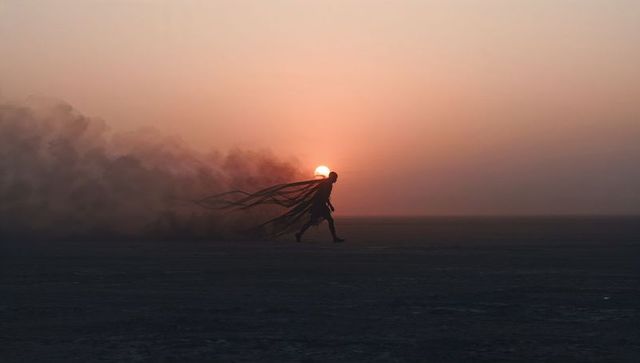 Walking silhouette at sunset trailing tattered cape and dust plume across desert dusk