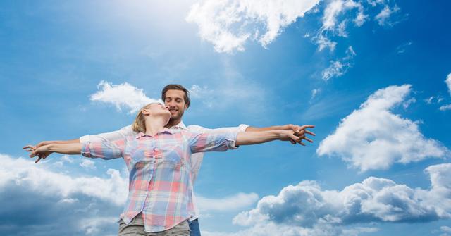 Loving couple standing with arms outstretched against a bright blue sky with fluffy clouds. Ideal for use in advertisements, relationship blogs, travel brochures, and lifestyle magazines to convey themes of love, freedom, and happiness.