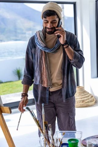 Male artist painting in home studio by waterfront talking on phone wearing beanie
