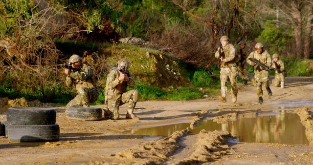 Soldiers participate in a military exercise, engaging in tactical maneuvers on a muddy dirt road surrounded by vegetation. They demonstrate teamwork and strategic movement with weapons in hand. This visual is ideal for use in content related to military training, defense strategies, teamwork development, and armed forces promotional materials.