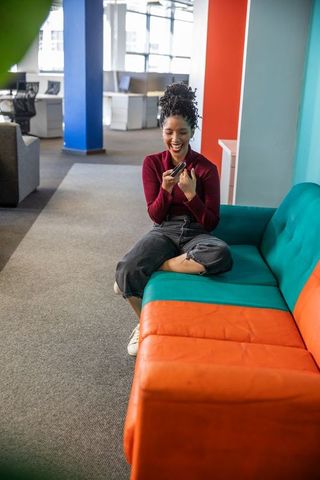Young african american woman laughing during phone call while relaxing on colorful office sofa
