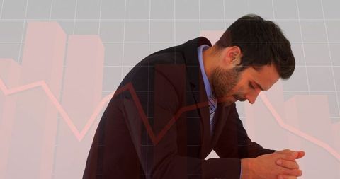 Stressed businessman leaning forward clasping hands with declining financial charts overlay