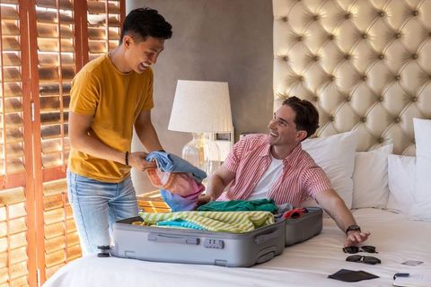 Diverse male friends laughing and packing suitcase on sunny hotel bed