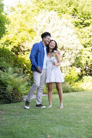 Laughing couple embracing in sunlit garden, woman holding white rose