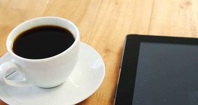 A cup of coffee sits next to a tablet on a wooden table, with copy space. This setup is typical for a modern workspace or a casual coffee break that includes digital interaction or reading.