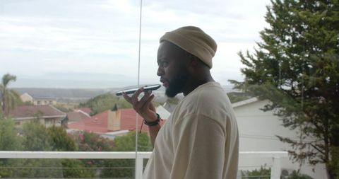 Man on balcony recording voice message on smartphone with overcast suburban view