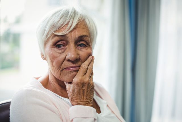 Elderly woman with white hair sitting indoors, looking thoughtful and concerned. Ideal for use in articles about aging, mental health, loneliness among seniors, or retirement planning.