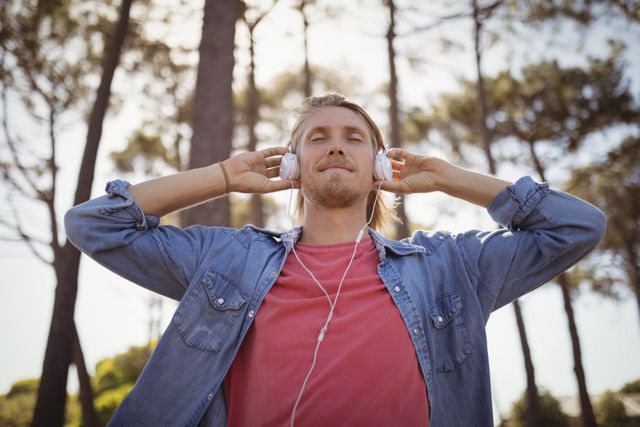 Young man standing in forest, wearing headphones, enjoying music. Ideal for concepts of relaxation, leisure, nature, and lifestyle. Suitable for use in advertisements, blogs, and articles about outdoor activities, mental health, and personal well-being.