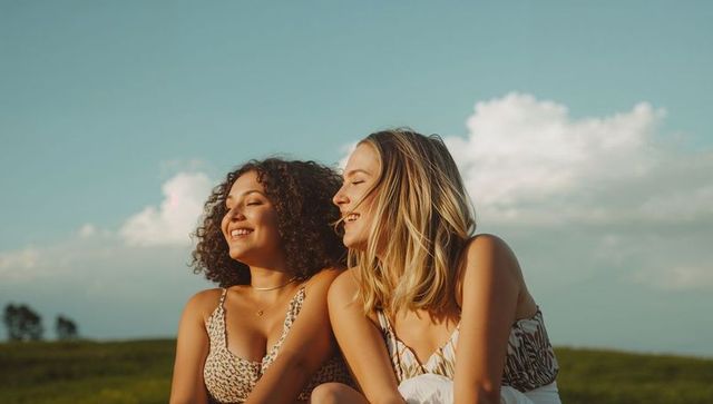 Two friends relaxing in sunlit meadow wearing patterned sundresses summer lifestyle