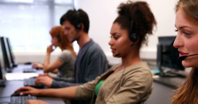 Group of call center agents from diverse backgrounds are seen working at computers and wearing headsets. The environment appears to be an office with good lighting. This image can be used for demonstrating teamwork, business communication, and customer service in a professional setting.
