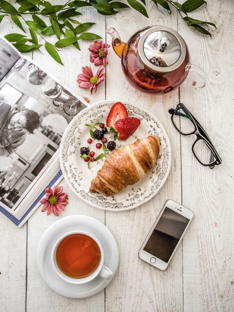 Elevated view of a charming breakfast spread featuring a croissant on a plate with berries and a sliced strawberry, accompanied by a cup of tea, a teapot, a smartphone, and glasses on a white wooden table. The setup is enhanced with a magazine, floral decorations, and green leaves. Perfect for promoting leisurely morning routines, food and beverage blogs, and lifestyle articles.