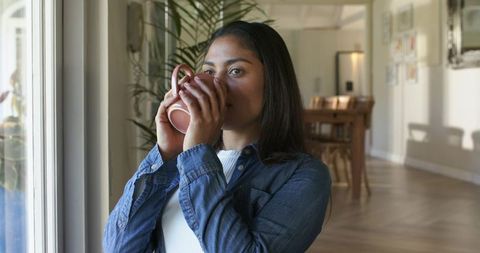 Sunlit morning at home sipping coffee by window, woman wearing denim shirt, cozy interior