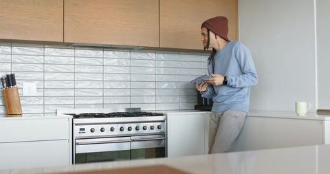 Mid-adult man standing holding tablet in modern minimal kitchen by stainless gas range