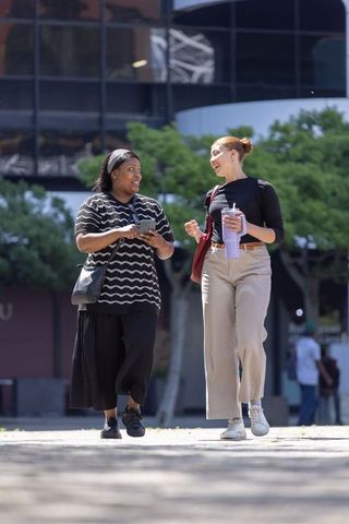 Two women walking and talking across urban plaza holding smartphone and reusable tumbler