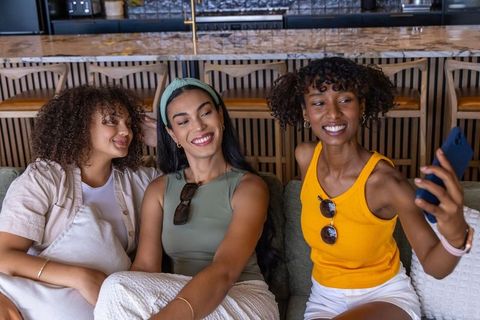 Three friends taking selfie on sofa in cozy cafe lounge, smiling and posing
