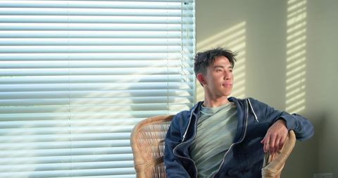 Asian man relaxing in wicker chair by sunlit blinds with striped shadows