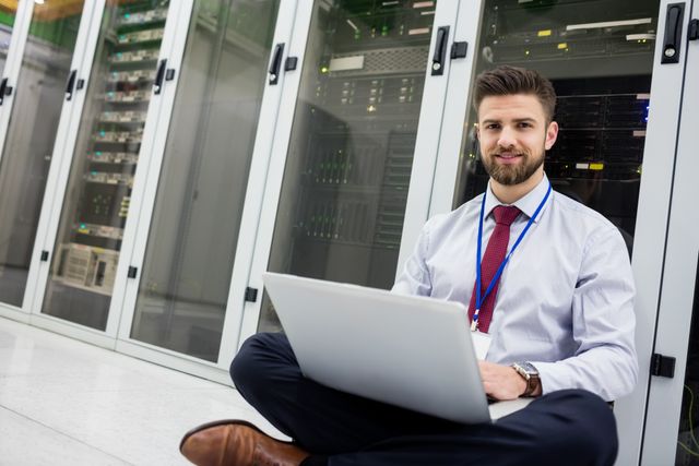 Technician sitting on floor using laptop in server room. Ideal for illustrating IT services, network management, data center operations, and professional technical support. Useful for websites, brochures, and articles related to technology, IT careers, and business infrastructure.