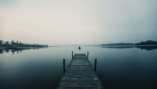 Wooden dock leading to lone rower on glassy lake at dawn quiet minimalist serene landscape