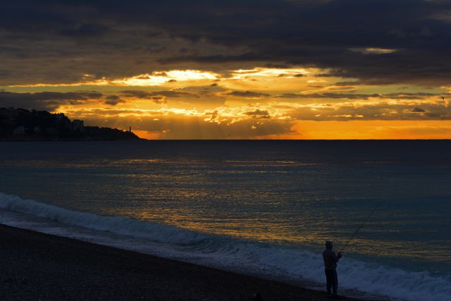 Silhouetted fisherman standing on beach fishing at sunset. Dark clouds and golden sky above calm sea and peaceful atmosphere. Suitable for themes related to nature, serenity, fishing, leisure, tranquil moments, and evening landscapes.