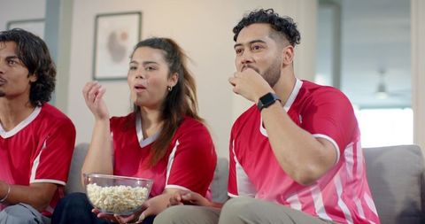 Multicultural friends wearing red jerseys sharing popcorn while watching soccer match