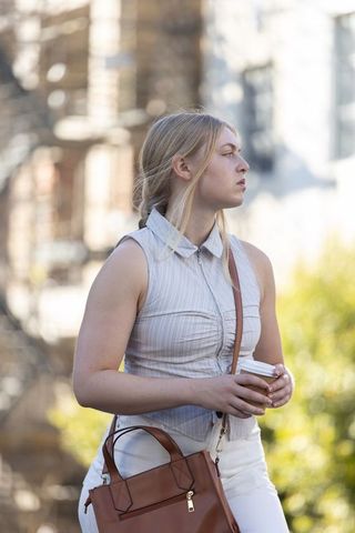 Young woman holding coffee cup on sunlit city sidewalk carrying brown leather handbag