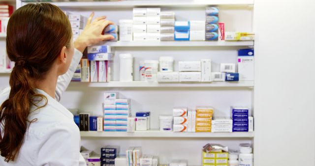 Female pharmacist organizing medication boxes on shelves in a modern pharmacy. She is wearing a lab coat and reaching for a medication package. Ideal for content related to pharmacies, medication management, healthcare services, and professional settings in the medical field.