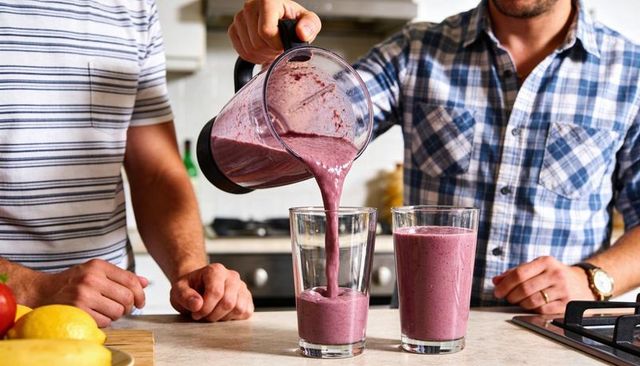 Two men pouring berry banana smoothie into glasses on kitchen counter, healthy morning routine
