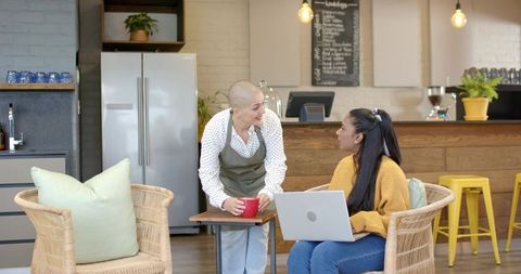 Barista chatting with indian customer holding red mug while customer typing on laptop