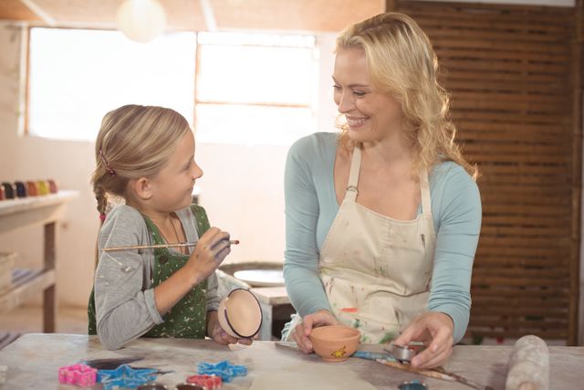 Female potter assisting young girl in painting pottery at a workshop. Both are smiling and engaged in the creative process. Ideal for use in educational materials, art class promotions, and articles about creative activities for children.