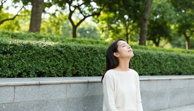 Young woman taking deep breath sitting on stone ledge in sunlit green park with hedge