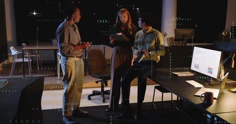 Night office team collaborating around tablet and monitor showing analytics charts after hours