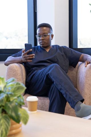 African male medical professional checking phone in clinic lounge wearing navy scrubs and glasses