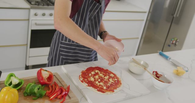 Person is preparing a homemade pizza in a modern kitchen, adding cheese as part of the cooking process. Various fresh ingredients including sliced levels of red and green bell peppers are arranged on the counter. Ideal for websites and blogs on cooking, culinary arts, or healthy homemade meals.