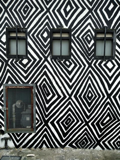 This image showcases a wall featuring a striking black and white geometric pattern, with three small windows and a vent. The bold design can represent modern architecture, street art, or urban settings. Ideal for use in articles on contemporary art, architectural features, or urban design inspirations. Great for backgrounds and textures in graphic design projects.
