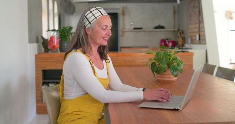 Mature woman typing on laptop at wooden kitchen table, working from home calmly with potted plant