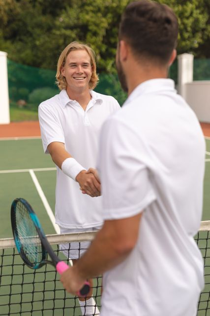 Two Caucasian men wearing tennis whites spending time on a court playing tennis on a sunny day, smiling and shaking hands over net, holding a tennis racket. Hobby sport leisure time.