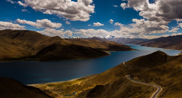Image shows a serpentine road winding alongside a tranquil lake, surrounded by a mountain range and a blue sky with scattered clouds. Perfect for travel websites, nature documentaries, or adventure tourism promotions depicting the beauty of remote and untouched wilderness landscapes.