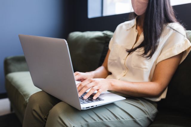 Midsection of asian businesswoman wearing face mask using laptop sitting couch. health and hygiene in workplace during coronavirus covid 19 pandemic.