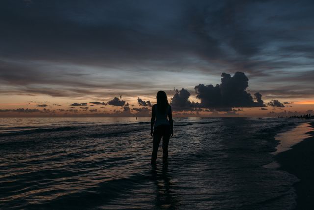 Shows silhouette of woman standing in shallow water at beach during vibrant sunset. Suitable for use in travel promotions, lifestyle blogs, relaxation and meditation content, and nature-inspired social media posts.