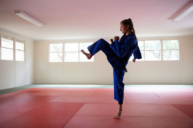 Caucasian female judoka wearing blue judogi, standing on a mat and warming up while preparing for her judo training in a bright studio.