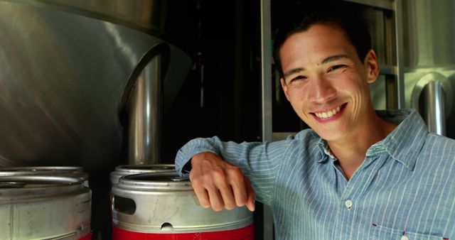 A young Asian man smiles while standing next to metal beer kegs, with copy space. His cheerful expression and casual attire suggest a relaxed environment, within a brewery or a storage area.