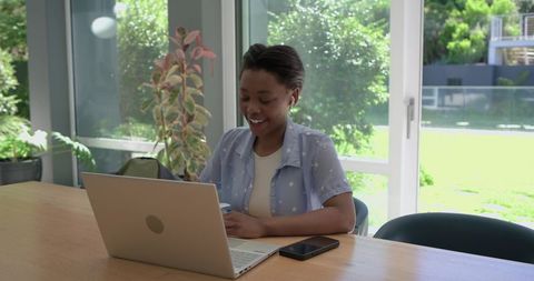 Young black woman working remotely on laptop and phone in bright home office with plants