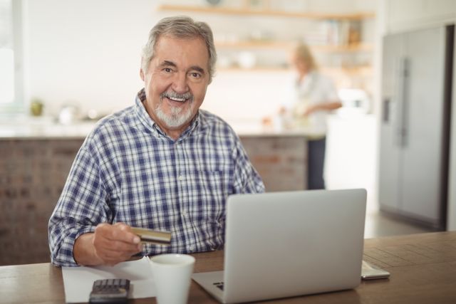 Smiling senior man paying bills online on laptop in kitchen - Download Free Stock Photos Pikwizard.com