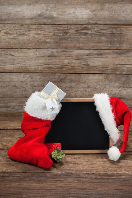 Santa hat and Christmas stocking with a blank chalkboard on a rustic wooden background. Ideal for holiday greetings, festive announcements, Christmas promotions, and seasonal messages.