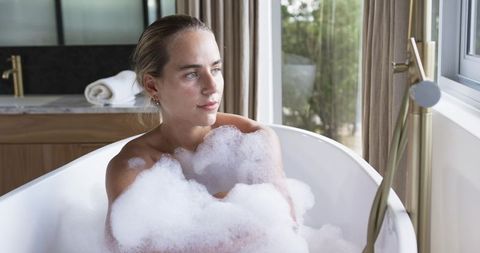 Woman relaxing in bubble bath by window in modern bathroom with brass faucet