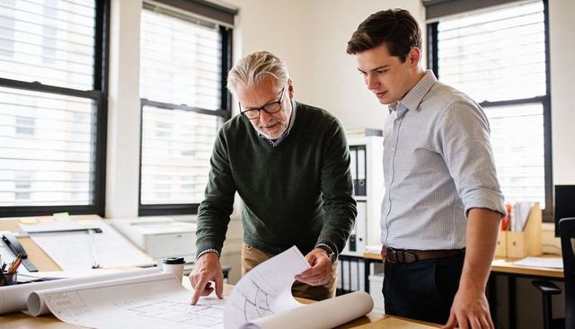 Senior architect mentoring young designer reviewing blueprints in light-filled studio