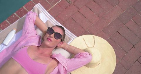 Poolside woman sunbathing on striped towel wearing pink bikini top and oversized sunhat