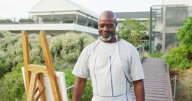 Senior African American man painting outdoors in a park while smiling. He is standing by an easel and canvas, wearing a casual gray shirt and apron. This image is suitable for creative hobbies, retirement activities, art therapy, elderly well-being, and active lifestyle promotions.