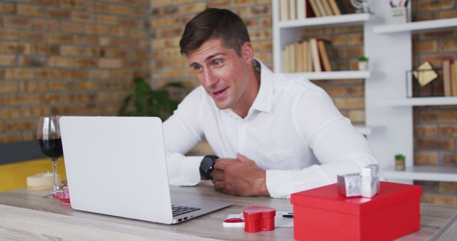 Smiling man having a video call on a laptop while a red gift box and romantic items are on the desk. Ideal for use in stories related to virtual dates, Valentine's Day celebrations, long-distance relationships, online meetings, and festive occasions.